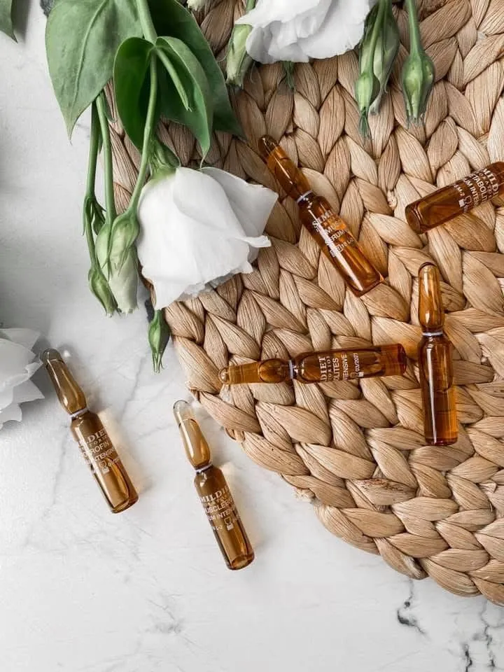 Cosmetic ampoules scattered on a woven rattan mat with white roses, marble background