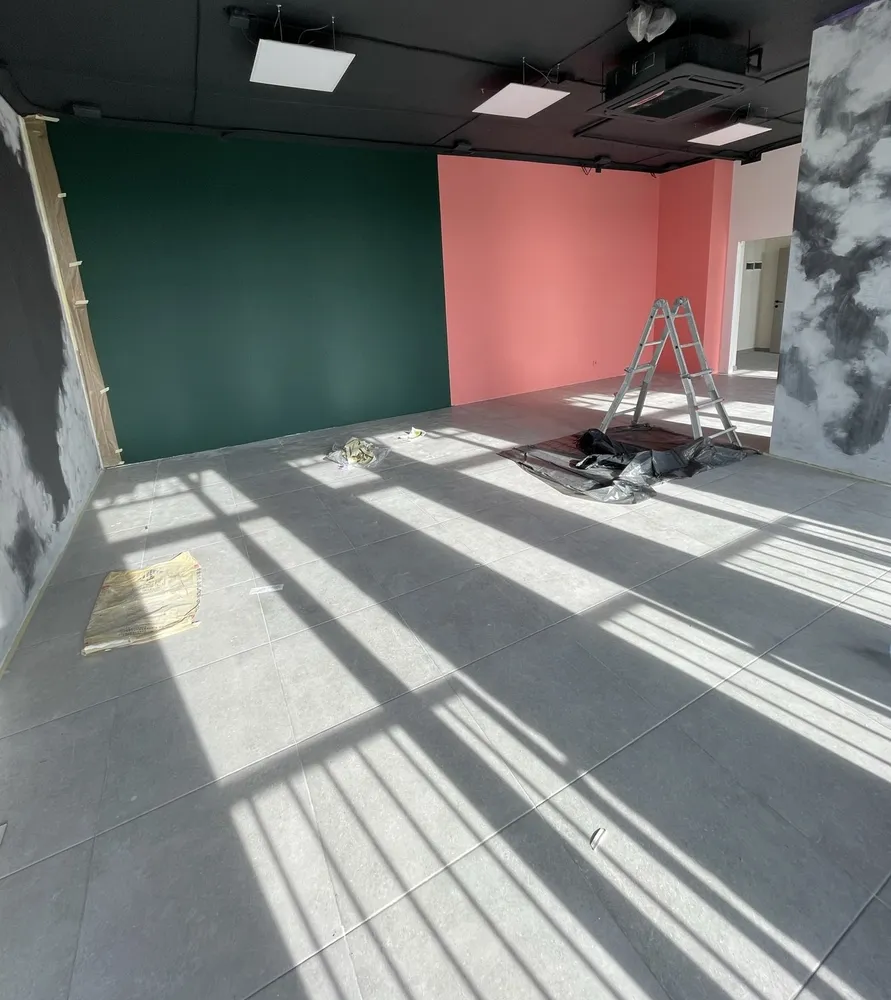 Empty studio under renovation: freshly painted green and pink walls, a stepladder, sunlight across a tiled floor